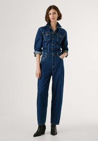 Denim jumpsuit med krage, knappelukking, smal nederst på ermene og vid benpassform. Har en mørkeblå farge og lys sømdetaljer.