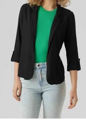 Frau trägt einen schwarzen Blazer über einem grünen Hemd kombiniert mit hellblauen Jeans vor einem einfarbigen hellgrauen Hintergrund.