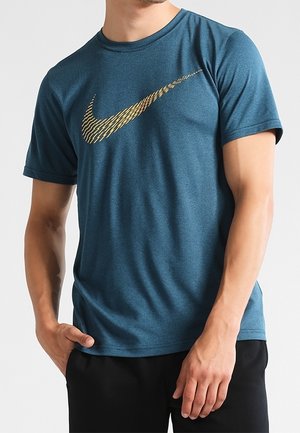 Mann trägt ein türkisfarbenes kurzärmeliges Sportshirt mit strukturiertem goldenem Nike-Swoosh-Logo, kombiniert mit schwarzen Hosen, steht mit der Hand in der Tasche.
