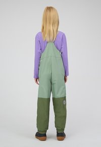 Grün-graue Latzhosen mit Latz-Design, verstärkten Knien und elastischen Bündchen, kombiniert mit einem langen lila Hemd darunter.