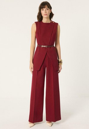 Kobieta w bordowym kombinezonie bez rękawów, z szerokimi nogawkami, paskiem w talii i złotymi bransoletkami, stojąca na jasnym, neutralnym tle.