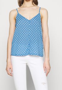 Femme portant un haut sans manches bleu à pois avec des boutons sur le devant et un jean skinny blanc déchiré, se tenant devant un fond uni.