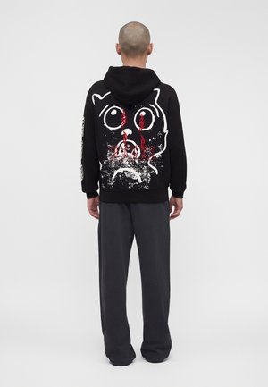 Mies, joka pitää mustaa hupparia, jossa on suuri kulunut surullisen kissan kasvot valkoisena ja punaisin kyynelin takana, yhdistetty tummanharmaisiin housuihin.