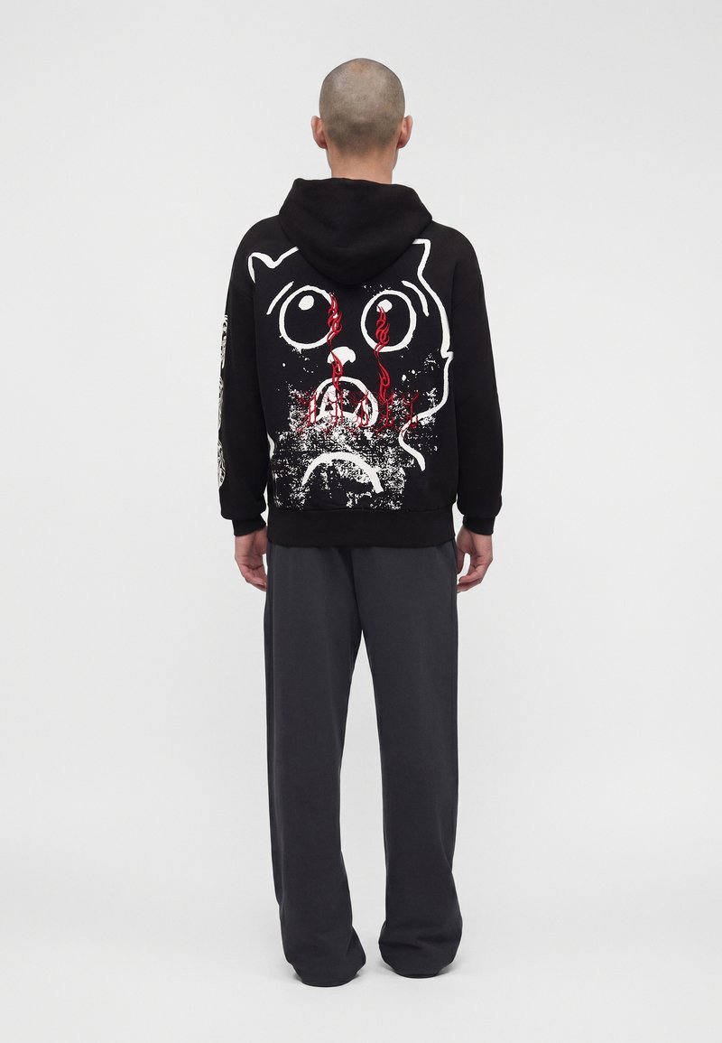 Hombre con sudadera negra con capucha con un gran gráfico desgastado de una cara de gato triste en blanco y lágrimas rojas en la espalda, combinado con pantalones gris oscuro.