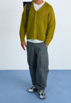Cardigan en laine vert avec des boutons, pantalon cargo gris et baskets noires et grises, porté avec un t-shirt gris et un sac à dos noir.