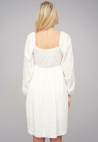 Vestido blanco con espalda cuadrada y mangas largas abullonadas. Presenta una falda fluida con tejido texturizado y un detalle de cintura ajustada.