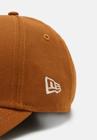 Gorra marrón hecha de tela texturizada, con un logo bordado en blanco y detalles de costura visibles. Ojete hundido en la parte superior para la ventilación.