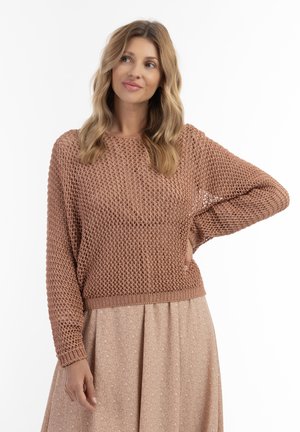 Kobieta z blond włosami, ubrane w luźny brązowy dzianinowy sweter i jasnobrązową spódnicę, stoi z jedną ręką na biodrze, patrząc na bok.