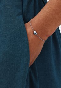 Arm in dark blue fabric pocket wearing a delicate silver chain bracelet with a small heart charm.