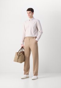 Beige stripe button-up shirt, beige trousers, and white sneakers. Holding a tan bag with brown accents and zipper detail.