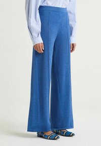 Pantalon à jambes larges en tissu bleu vif à texture lisse. Il présente une taille haute et un léger éclat, associé à des chaussures à motifs.