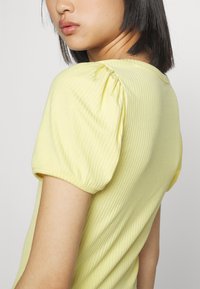 T-shirt amarelo com nervuras, com mangas curtas e bufantes, design justo e textura subtil. Detalhes de costura visíveis nos ombros e no decote.