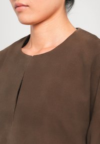 Gros plan du bas du visage et du cou d'une personne portant un haut brown à encolure ronde, sur fond uni.