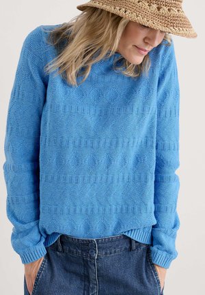 Pull en laine bleu avec un motif zigzag texturé, col rond et poignets côtelés. Associé à un pantalon en denim, mettant en avant une coupe décontractée.