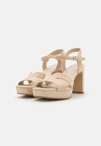 Beige suede platform sandals with a chunky heel, featuring a crossed strap design and a buckle fastener. Smooth sole and open toe.
