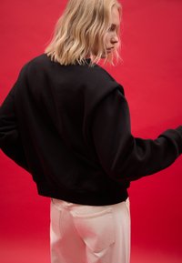 Felpa nera corta con scollo rotondo e polsini a coste, abbinata a pantaloni chiari su uno sfondo rosso vivace.