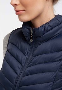 Marineblauw gewatteerd bodywarmer van glanzend, doorgestikt materiaal met ritssluiting en hoge kraag. Voorzien van schuine naden en zichtbare stikseldetails.