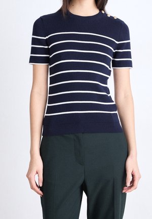 Femme portant un pull bleu marine à manches courtes avec des rayures blanches horizontales et des boutons dorés sur l'épaule gauche, assorti à un pantalon foncé.