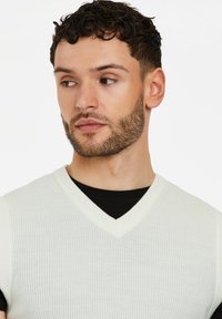 Ärmelloser V-Ausschnitt-Pullover aus leichtem Cremefarben mit Rippstruktur, über einem schwarzen Hemd getragen, mit feiner Textur und dezenten Details am Ausschnitt.