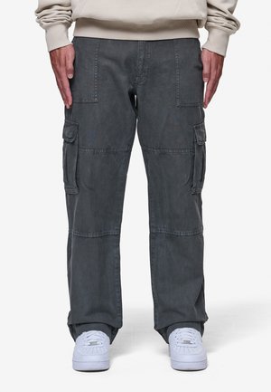 Homme portant un pantalon cargo gris foncé avec plusieurs poches et des baskets blanches, debout les bras détendus le long du corps.