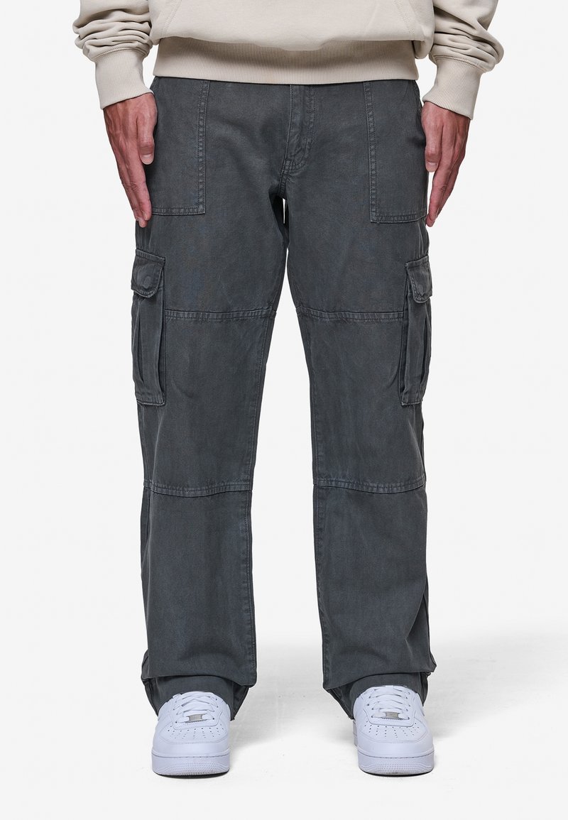 Homme portant un pantalon cargo gris foncé avec plusieurs poches et des baskets blanches, debout les bras détendus le long du corps.