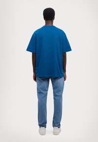 Blå bomull t-skjorte med korte ermer og rund hals, sammen med lyseblå jeans og hvite joggesko, sett fra baksiden.