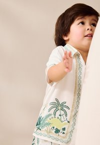 Balta trumpomis rankovėmis marškinėliai su žaliu ir geltonu dinozaurų bei augalų raštu, su geometriniu akcentu palei sagų juostelę.