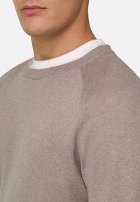 Gros plan d'une personne portant un pull en tricot beige superposé à un t-shirt blanc à col rond, montrant les détails du cou et des épaules.