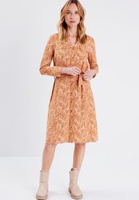 Langärmliges Kleid aus weichem orangenem Stoff mit floralem Muster, einer Knopfleiste vorne und Taillenschnürung, kombiniert mit hellen Knöchelstiefeln.