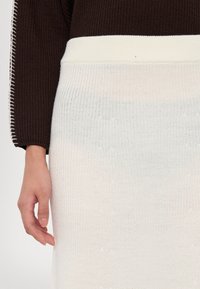 Stickad tröja med mörkbrun övre del och vit nedre del, med strukturerat mönster och en vit ribbad midja. Hand synlig.
