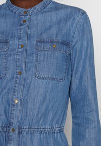 Vaaleansininen farkkumekko, jossa on korkea kaulus, kaksi rintataskua, kultaiset painonapit ja joustava vyötärö istuvaa siluettia varten.