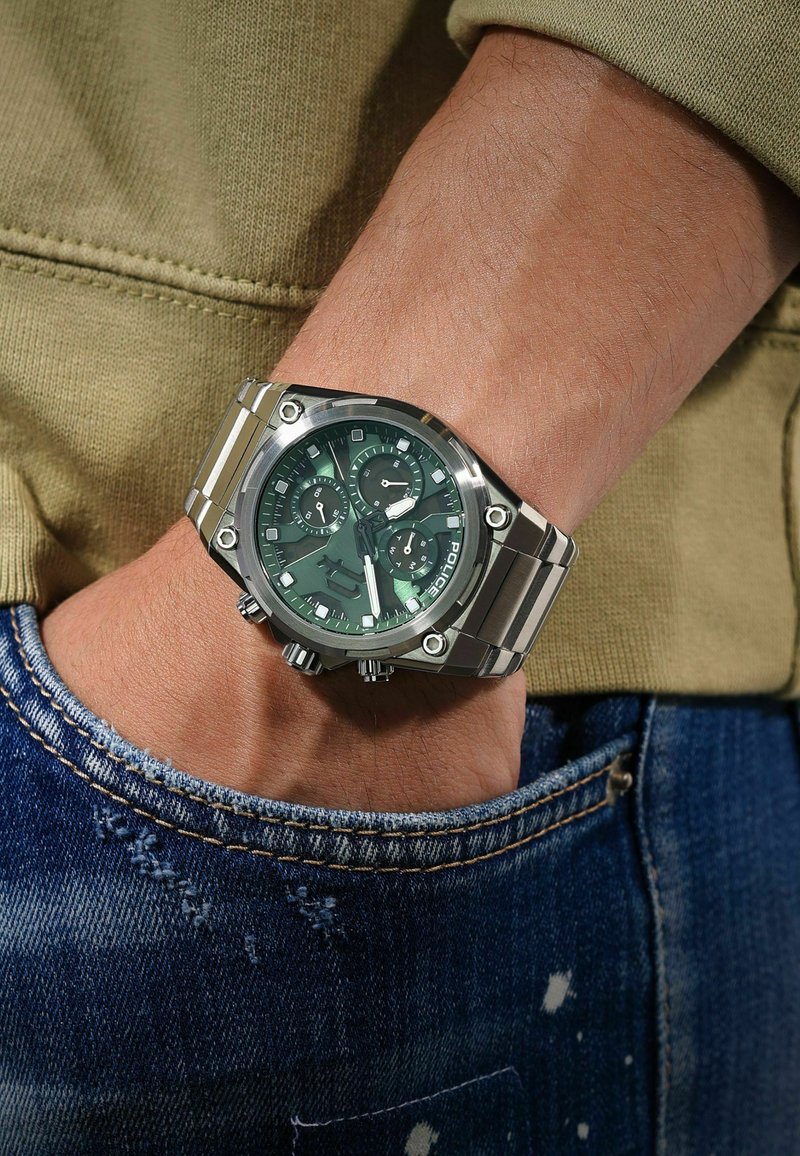 Zilveren roestvrijstalen polshorloge met groene wijzerplaat, subdials en gegraveerde accenten, gedragen op een hand boven denim stof.