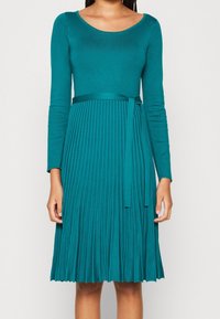 Robe longue teal à manches longues avec encolure arrondie, corsage ajusté et jupe plissée. Comprend une ceinture en tissu assortie à la taille pour un détail supplémentaire.