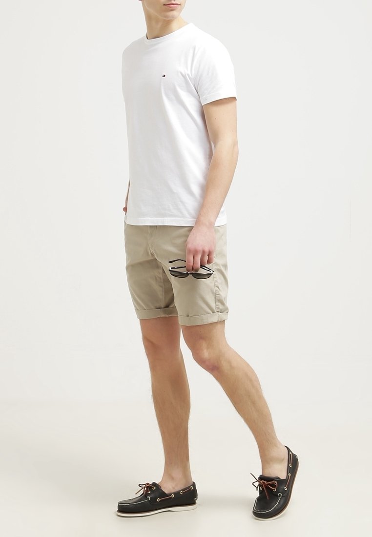 Homme portant un t-shirt blanc, un short beige et des chaussures bateau noires tenant des lunettes de soleil, marchant sur un fond clair uni.