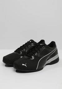 Des chaussures de sport noires avec un dessus en synthétique lisse, des accents texturés et un logo blanc. Elles possèdent une semelle rembourrée et un design à lacets.