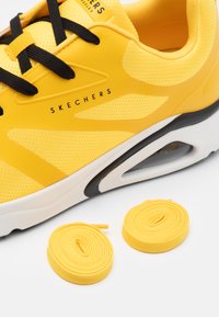 Sneaker Skechers gialla con tomaia in mesh, lacci neri, sovrapposizioni texturate e intersuola bianca imbottita. Include lacci gialli coordinati.