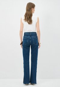 Jeans acampanados de cintura alta en denim azul oscuro, con bolsillos traseros y detalles de botones a lo largo de las costuras laterales. Textura suave, corte ajustado.