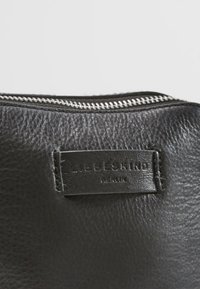 Czarna skórzana torba z fakturowaną powierzchnią, wyposażona w srebrny zamek błyskawiczny i prostokątną, wytłaczaną etykietę marki.