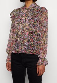 Bloes met bloemenprint van doorschijnende zwarte stof met roze, paarse en gele bloemen. Beschikt over een gerimpelde kraag en mouwen met elastische manchetten.