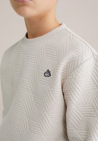 Világosszürke pulóver hatszögletű mintával és bordázott nyakkivágással, a mellrészen kis fekete hímzett logóval.