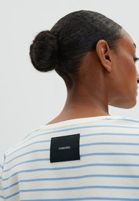 Gestreiftes T-Shirt in Creme mit hellblauen Streifen, mit einem schwarzen Stoffetikett auf der Rückseite, das "Eines Tages" besagt. Glatte Textur und lässiges Design.