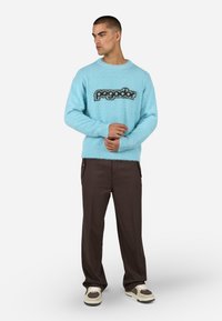 Šviesiai mėlynas pūkuotas megztinis su juodu „pagador“ tekstu ant krūtinės, derinamas su tamsiomis kelnėmis ir smėlio spalvos sportbačiais. Patogi tekstūra ir laisvas kirpimas.