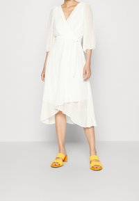 Vit omslagklänning med prickigt mönster, V-ringning och genomskinliga puffärmar. I kombination med gula blockklacksandaler. Asymmetrisk fåll.