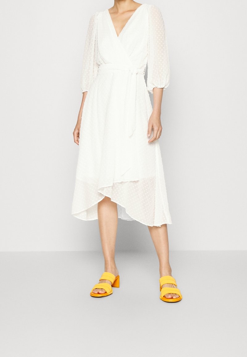 Vit omslagklänning med prickigt mönster, V-ringning och genomskinliga puffärmar. I kombination med gula blockklacksandaler. Asymmetrisk fåll.