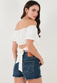 Hvid off-shoulder bluse med flæser på ærmerne, bundet bagpå. Sammen med blå denimshorts med frynset kant og mærke.