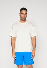 Beżowa koszulka z krótkim rękawem, z napisem "CFC" w kolorze niebieskim oraz logo Nike w kształcie łyżwy. Noszona z niebieskimi szortami sportowymi. Gładkie tło.