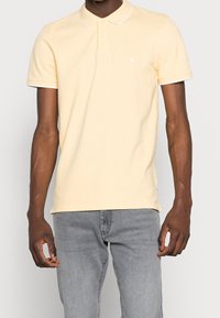 Pikétröja i mjuk gul bomull med vit krage och knappslå. Har en liten logotyp på bröstet och korta ärmar. Matcher med grå jeans.