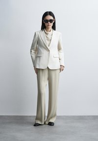 Kvinde med glat sort hår iført sorte solbriller, cremefarvet blazer, matchende vidde bukser og sorte sko, stående mod en ensartet lys baggrund.