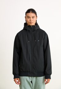 Black zip-up jacket with a hood, featuring drawstrings, ribbed cuffs, and a logo on the chest. Smooth fabric with a relaxed fit.