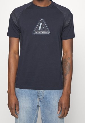 Mann trägt ein marineblaues Emporio Armani T-Shirt mit einem dreieckigen Logo und hellblaue Jeans, sichtbares Tattoo auf dem rechten Unterarm.